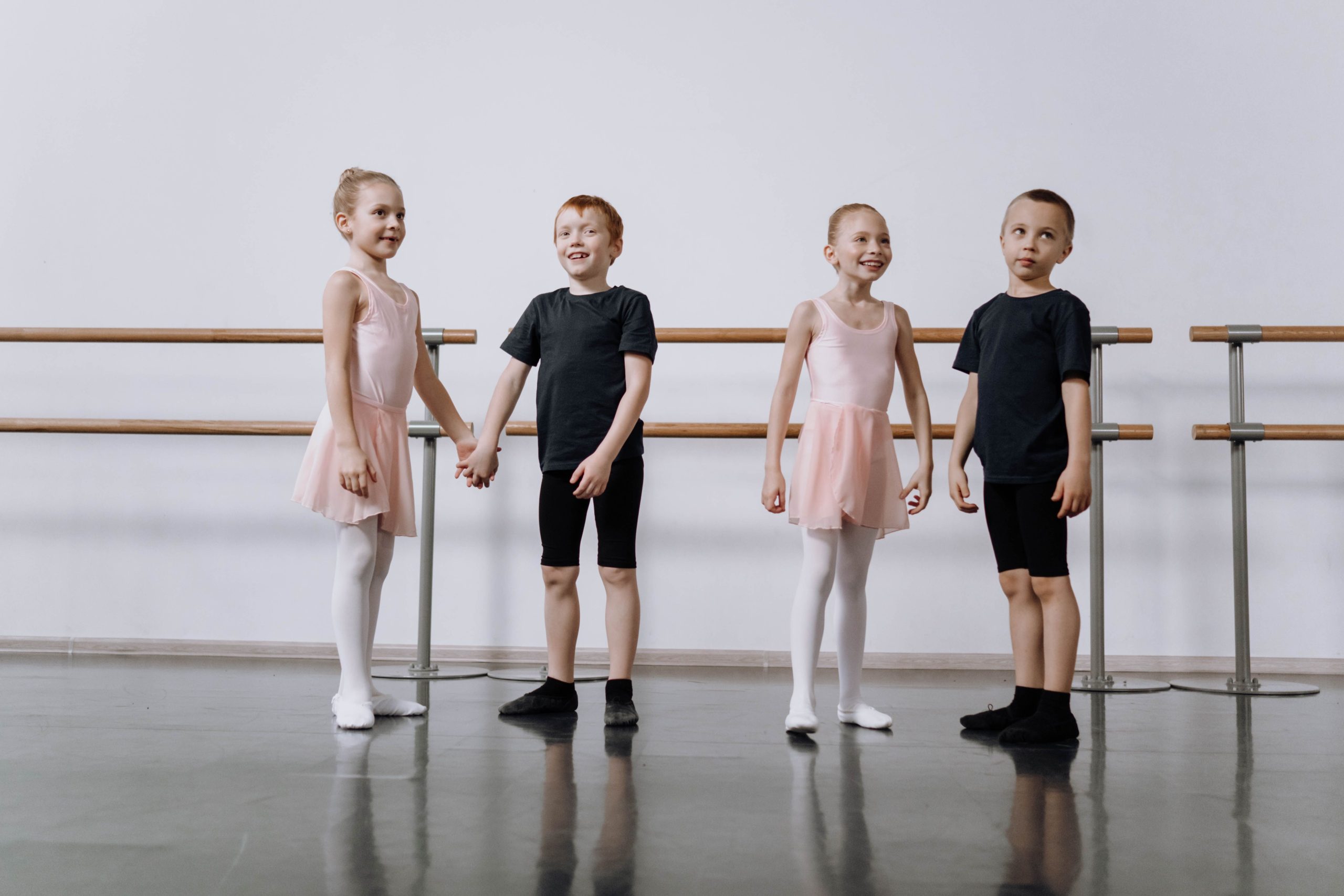 Four young children in ballet attire practice dance in a bright studio setting.